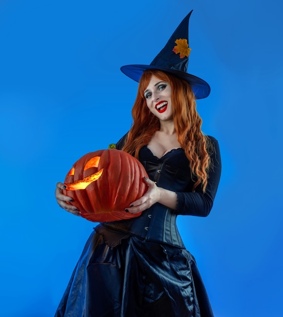 Фото Ведьма на хэллоуин женщина с длинными красными волосами и праздничным макияжем в шляпе темной конической ведьмы. красавица-готическая девушка с резной тыквой в арт-дизайне party celebrating all saints'â