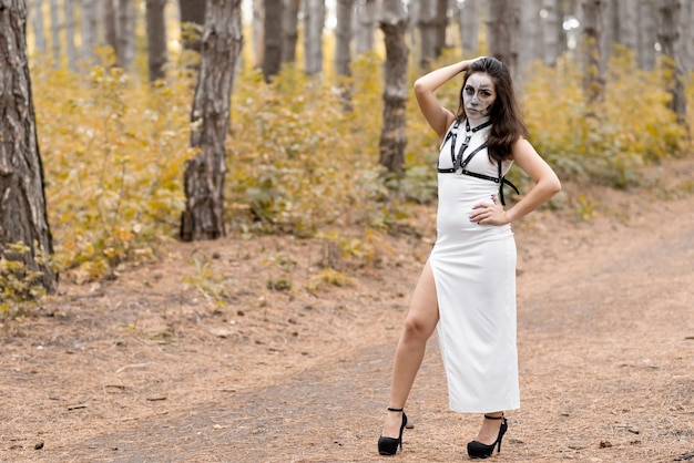 Halloween young beautiful girl in a white dress with makeup on her face Posing in the autumn forest Skeleton face makeup The day of the Dead