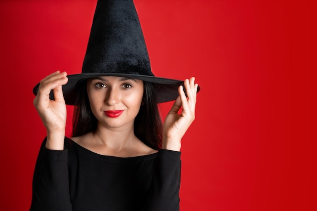 Halloween A young beautiful woman in a witch39s hat and a black dress on a red colored background Space for text