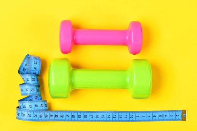 Halters in roze en lichtgroene kleuren en meetlint leggen top op gele achtergrond. Centimeter in blauw in de buurt van kleurrijke sportuitrusting. Gewichtsverlies en sportconcept. Gym en gezond leven tools.