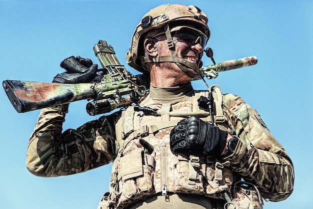 Halve lengte lage hoek locatie shot van special forces soldaat in velduniformen met wapens, portret op blauwe hemelachtergrond