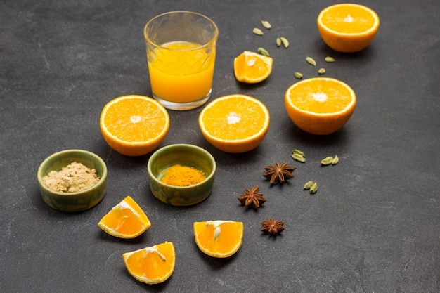 Halves of orange, star anise and cardamom on table. Turmeric and ginger in green bowl. Glass of juice. Black background. Top view.