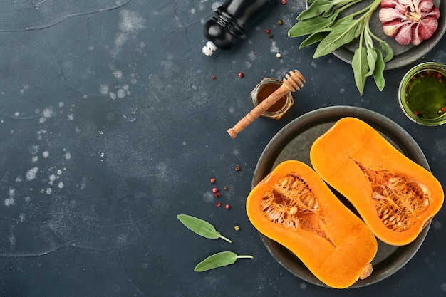 Metà della zucca butternut organica cruda con foglia di salvia, aglio pepe multicolore, miele, sale e pepe su sfondo nero di ardesia, pietra o cemento. sfondo di cibo. vista dall'alto con copia spazio.