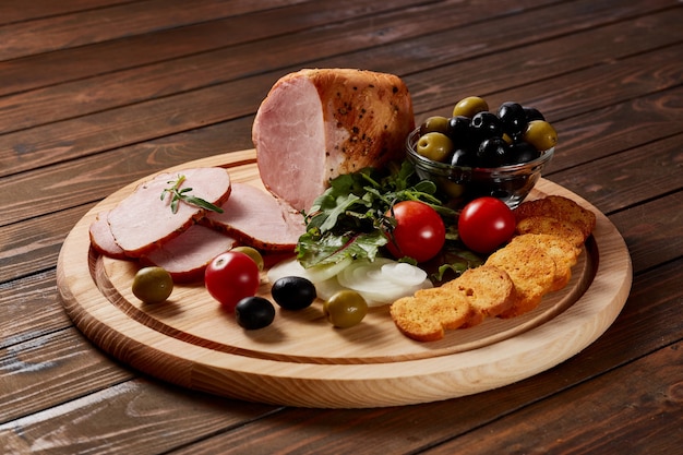 Ham garnished with vegetables, fruits and herbs with sauces and bread on a wooden plate on a dark wooden table.