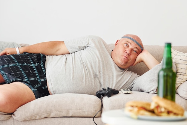 Hamburgers and beer man lying on the sofa funny overweight man
in casual clothes is indoors at home