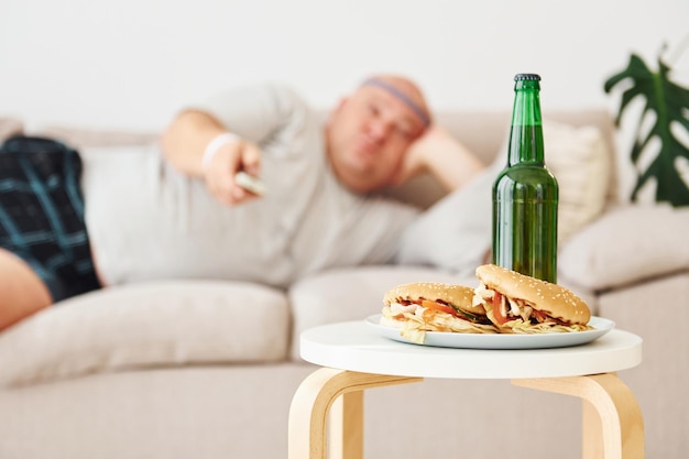 Hamburgers and beer man lying on the sofa funny overweight man\
in casual clothes is indoors at home