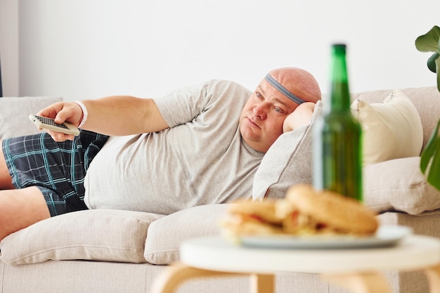 Hamburgers and beer Man lying on the sofa Funny overweight man in casual clothes is indoors at home