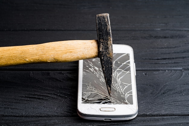 Hammer brokes modern sensory smartphone isolated dark . Expensive cellphone smashed by big hammer on wooden surface.