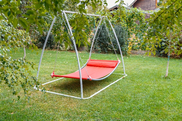 Hammock swing in metal frame with nobody on green lawn in backyard near house cottage. Rest relax relaxation alone on Red hammock swing in Summer garden.