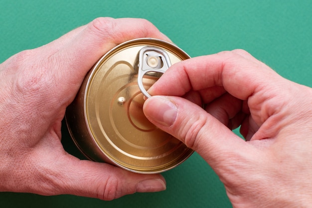 The hand of an adult holds a can of canned food, the second hand pulls the key, opens the can. Top view on green background with copy space. Close-up