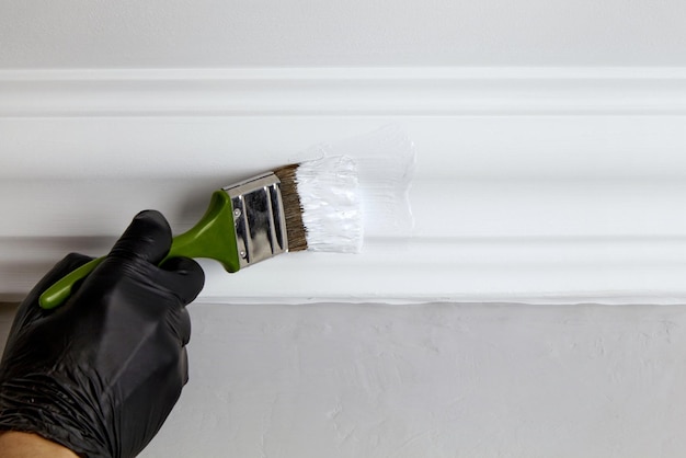 A hand in a black rubber glove with a brush paints the ceiling plinth with white paint Repair in the room