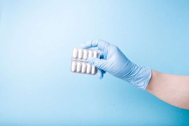 Hand in a blue disposable medical glove holds a blister with white capsules on blue surface