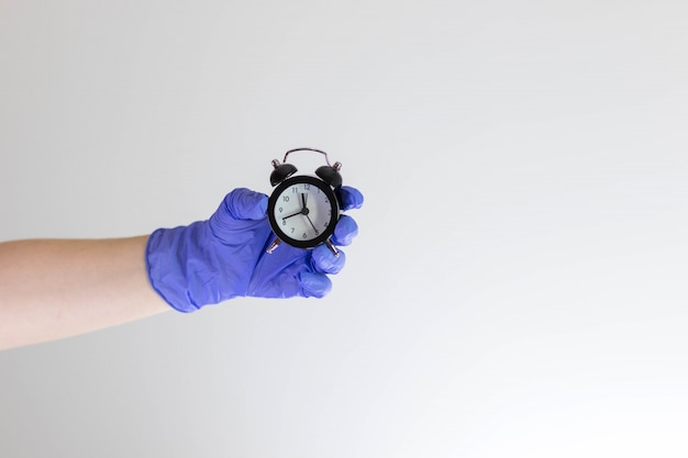 Hand in blue medical glove holding small alarm clock.