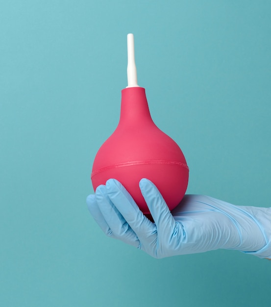 A hand in a blue medical glove holds a pink rubber enema on a blue background