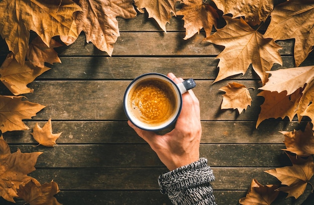 Hand of a caucasian male holding a hot coffee cup on an aged wooden table with autumn leaves around