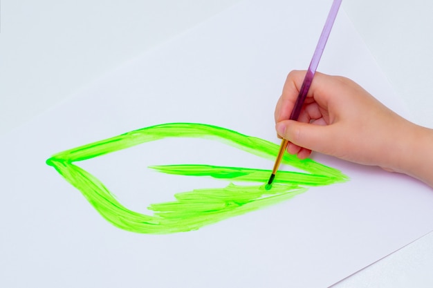 Hand of child drawing green leaf with brush by watercolors on white paper. Earth day concept.