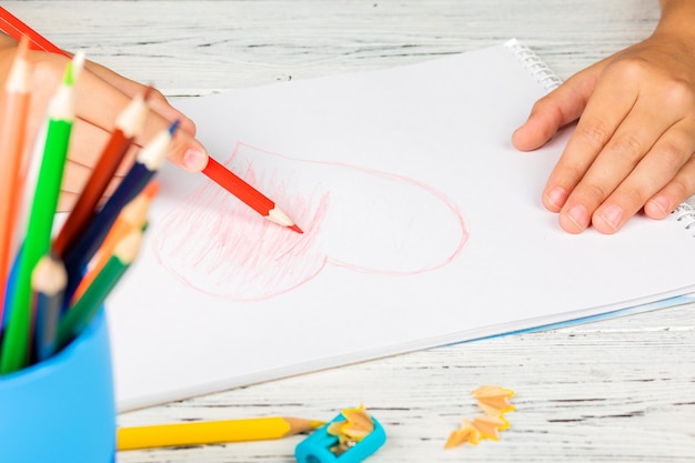 Foto mano di bambini che disegnano cuore rosso con matita colorata su carta bianca su tavola di legno