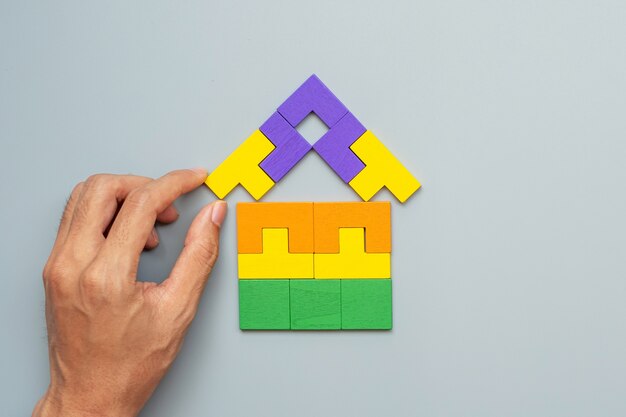 Hand connecting Home shape block with colorful wood puzzle pieces on gray