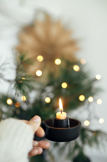 Hand in cozy sweater holding burning candle on background of\
lights in scandinavian room hygge home