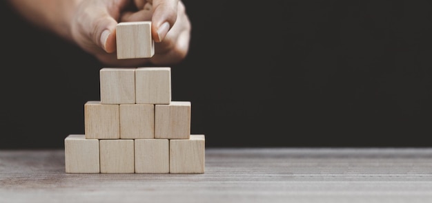 Hand die een houten kubus bovenop een houten blok zet die stapelt als een stapsgewijze groei van het bedrijf naar succes