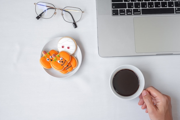 Hand die een koffiekopje vasthoudt en Halloween-koekjes eet tijdens het gebruik van een computerlaptop. Happy Halloween, online winkelen, Hallo oktober, herfstherfst, Feestelijk, feest- en vakantieconcept