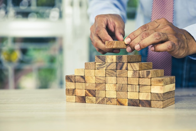 Hand die houten bloktorenstapel in piramidetrap plaatst met voorzichtigheid om instorting of neerstortingsconcepten van financieel risicobeheer en strategische planning en bedrijfsuitdagingsplan te voorkomen