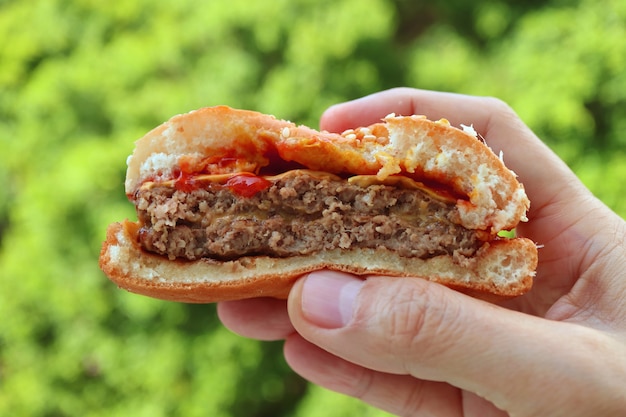 Hand die in halve hamburger met onscherp groen gebladerte wordt gespleten