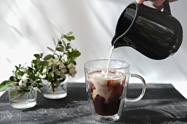 hand die melker vasthoudt en melk in een koffiemok giet koude koffie met ijsblokjes en bloemen op een grijze tafel ochtend ontbijt tijd ontspannen concept