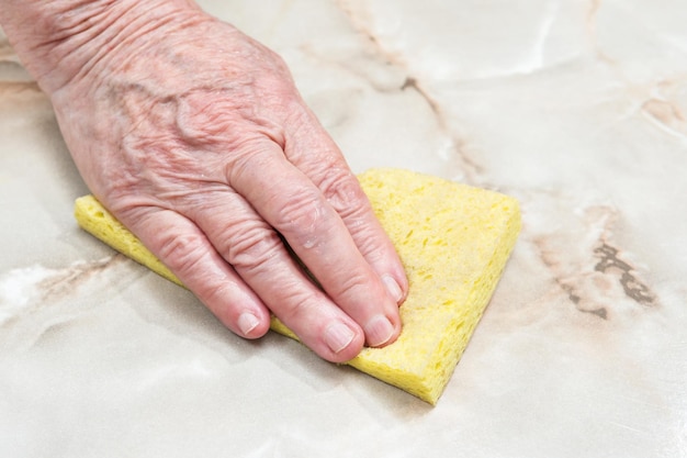 Hand of an elderly woman wipes the table dry with a yellow sponge