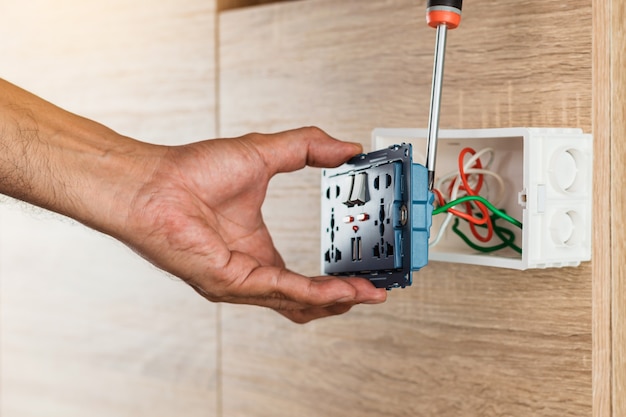 Hand of an electrician is using a screwdriver to attach the\
wires to the universal wall outlet ac power plug with usb port and\
on-off in a plastic box on a wooden wall.