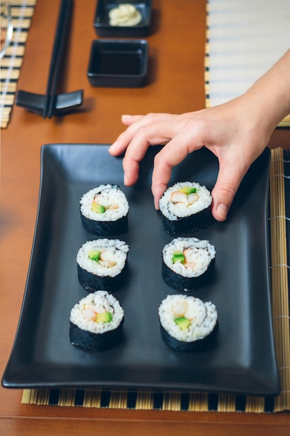 黒の長方形のトレイの上の海苔シートにご飯、アボカド、エビと日本の巻き寿司を置く女性シェフの手