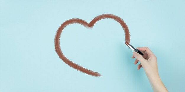 Mano di donna disegnare parte del cuore con rossetto marrone su sfondo blu.