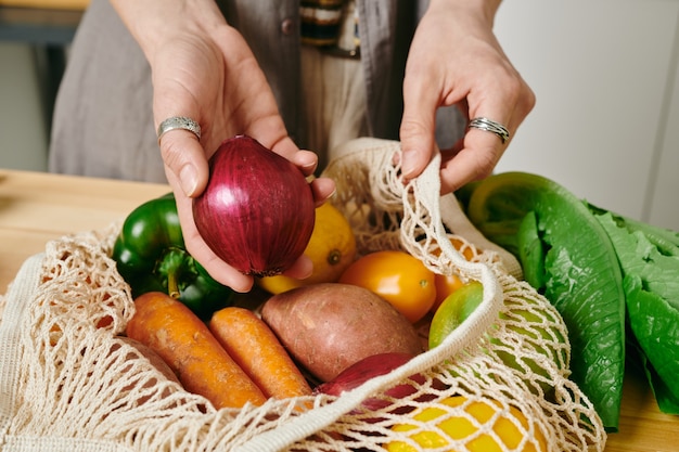 他の野菜と一緒にバッグの上に新鮮なタマネギを保持している女性の手