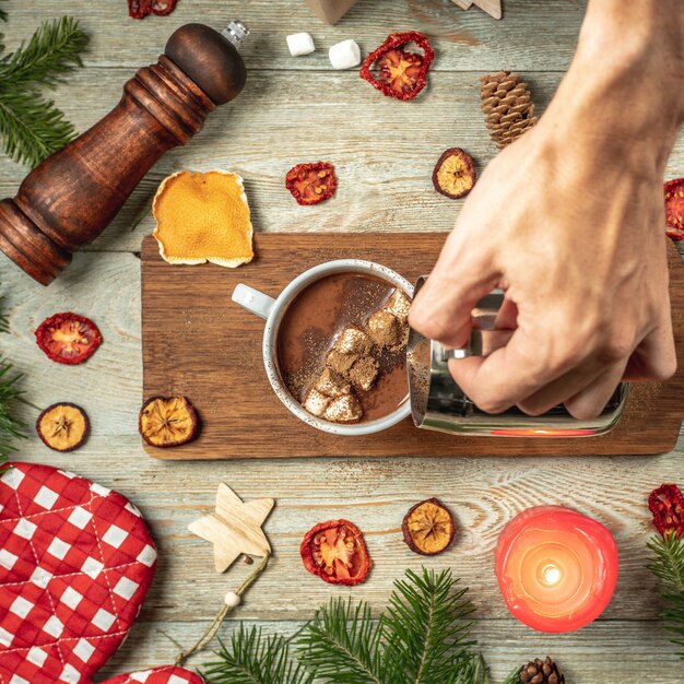 Hand giet warme chocolademelk in een witte kop. Rondom op een houten tafel staan feestelijke accessoires. Concept van gezellige kerst- en nieuwjaarsstemming.