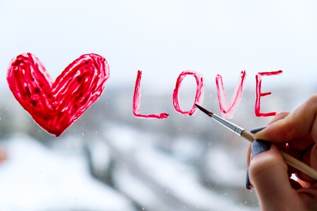 Foto mano di una ragazza con un pennello che dipinge un cuore rosso amore su una finestra, resta a casa, tempo libero in quarantena, stiamo tutti bene, san valentino, amore, romanticismo