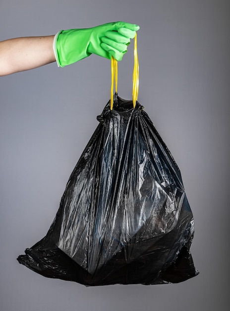 Hand in glove holding handles of black plastic disposable trash bag full of garbage waste on green banner background with copy space for text