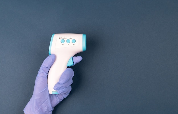 Hand in glove holding ir thermometer on dark blue background.  Distant temperature check.