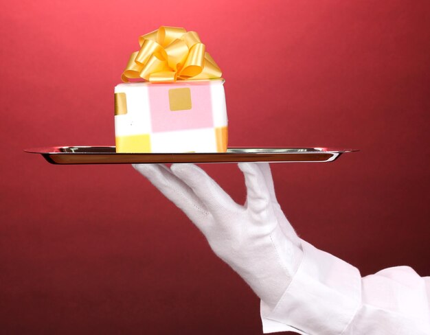 Hand in glove holding silver tray with giftbox on red background