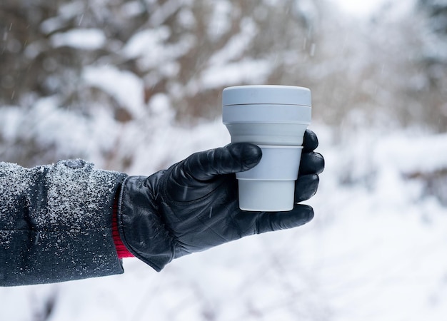 Hand in glove holding take away eco silicone coffee cup take out tea mug in winter in snowy weather outdoors