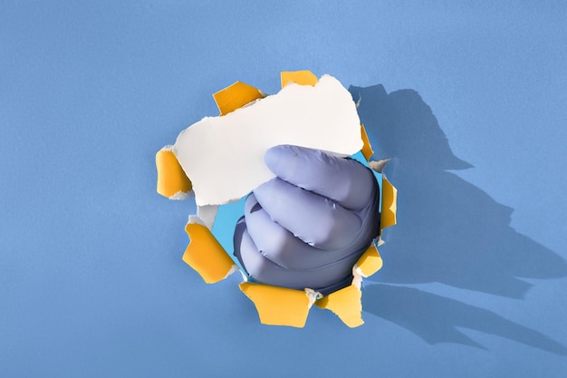 Hand in glove through paper hole holding torn piece of paper copyspace place for text on blue paper