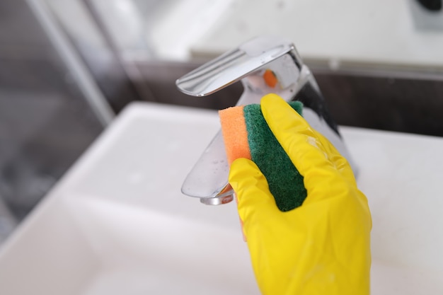 A hand in a glove washes the faucet in the bathroom with a sponge