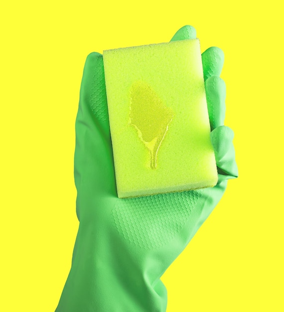 Hand in green glove holding cleaning sponge with liquid fluid detergent drop for washing on yellow background