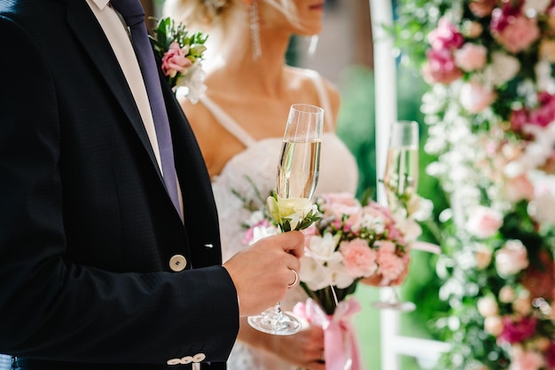 The hand groom's holds a glass of champagne on the background of the bride under the arch decorated with flowers and greenery in the backyard banquet area couple newlyweds Wedding ceremony