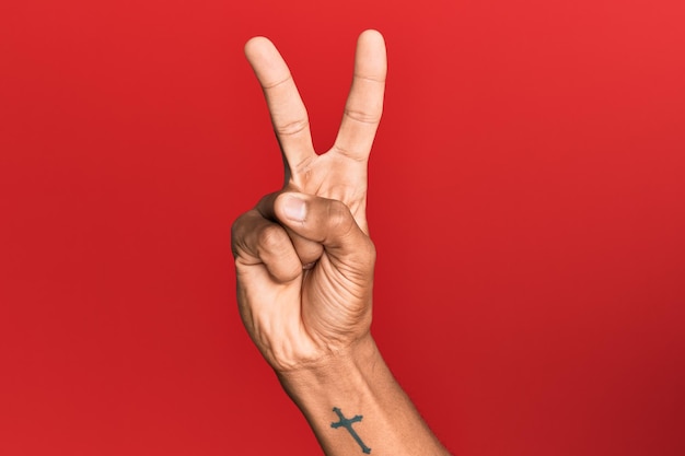 Hand of hispanic man over red isolated background counting number 2 showing two fingers, gesturing victory and winner symbol