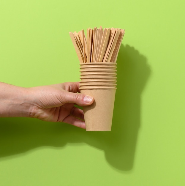 Hand hold brown paper cardboard cups and wooden stirring sticks on a green background. Eco-friendly tableware