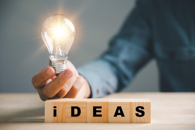 Фото Рука держит светящуюся лампочку со словом «идеи» на деревянном блоке. представляет концепцию новых идей, инноваций, вдохновения в бизнес-стартапе. символизирует способность мыслить творчески и развивать успех.