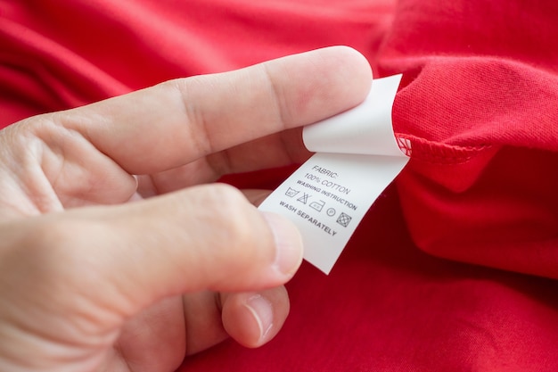 Hand hold and reading at white laundry care washing instructions clothes label on red cotton shirt