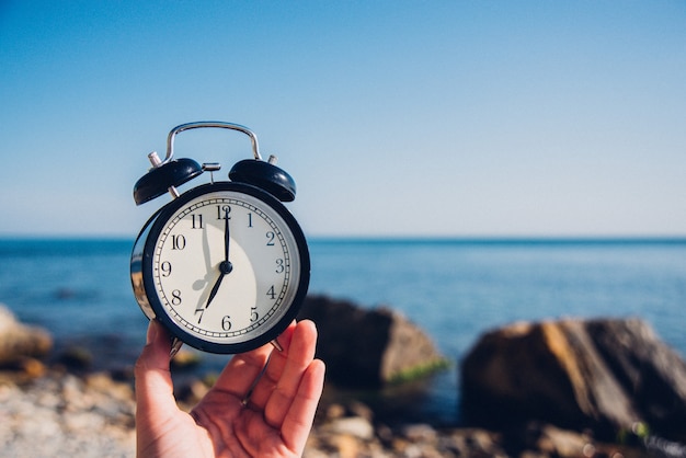 Foto tenere in mano l'orologio sullo sfondo della spiaggia. sveglia sul mare e la sabbia sullo sfondo in tempo diverso con le vacanze estive. concetto di tempo di vacanza.