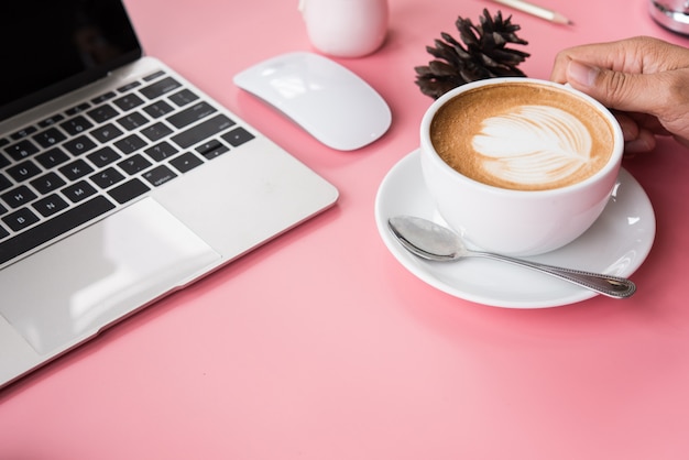 写真 ノートパソコンでピンクの机の上にコーヒーを1杯持っている手。