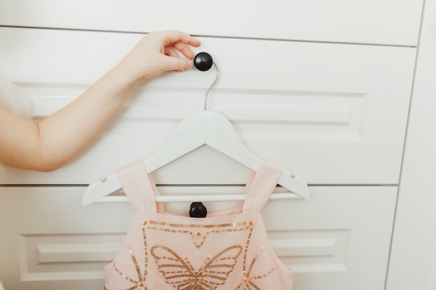 Photo hand holding beautiful dressy lush pink dress for girls on hanger at the background of wardrobe